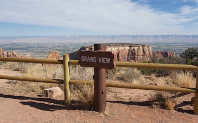 A scenic overlook featuring a sign labeled 'Grand View' situated next to a wooden fence. In the background, expansive rocky mesas and a wide valley stretch towards a distant mountain range under a partly cloudy sky.