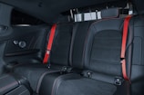 The image features the back seat of a car interior, showcasing black leather upholstery with red stitching and red seat belts. The design is sleek and modern, indicating a focus on style and comfort.