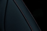 A close-up view of a textured dark material featuring a prominent seam with detailed stitching. The material appears to be a type of leather or similar fabric, characterized by fine, dark-colored stitches running along a curved seam.