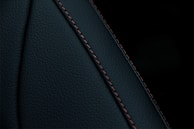 A close-up view of a textured dark material featuring a prominent seam with detailed stitching. The material appears to be a type of leather or similar fabric, characterized by fine, dark-colored stitches running along a curved seam.