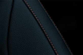 A close-up view of a textured dark material featuring a prominent seam with detailed stitching. The material appears to be a type of leather or similar fabric, characterized by fine, dark-colored stitches running along a curved seam.