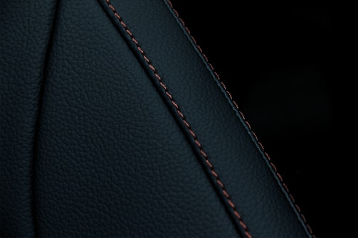 A close-up view of a textured dark material featuring a prominent seam with detailed stitching. The material appears to be a type of leather or similar fabric, characterized by fine, dark-colored stitches running along a curved seam.