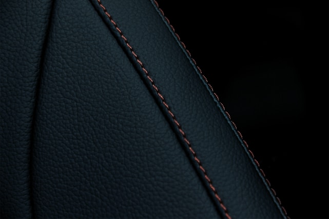 A close-up view of a textured dark material featuring a prominent seam with detailed stitching. The material appears to be a type of leather or similar fabric, characterized by fine, dark-colored stitches running along a curved seam.