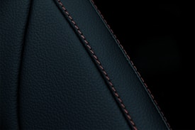 A close-up view of a textured dark material featuring a prominent seam with detailed stitching. The material appears to be a type of leather or similar fabric, characterized by fine, dark-colored stitches running along a curved seam.