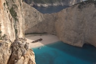 A secluded beach is surrounded by towering limestone cliffs. The sandy shoreline features a shipwreck, creating a striking centerpiece. The turquoise blue water is vibrant and contrasts with the pale colors of the cliffs and sand. Large cliffs enclose the bay, providing an isolated and tranquil setting.