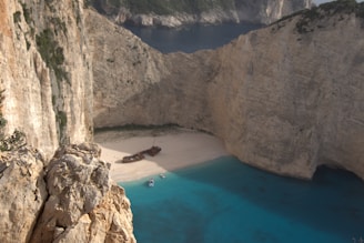 A secluded beach is surrounded by towering limestone cliffs. The sandy shoreline features a shipwreck, creating a striking centerpiece. The turquoise blue water is vibrant and contrasts with the pale colors of the cliffs and sand. Large cliffs enclose the bay, providing an isolated and tranquil setting.
