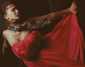 A person is dressed in a flowing red gown with a striped scarf and blue beaded necklace. The composition evokes a sense of elegance and movement.