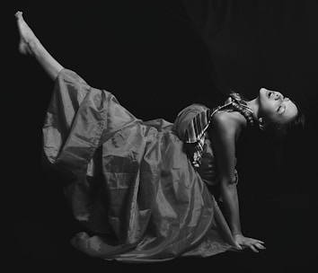 A black and white photograph of a person in a dramatic pose, wearing a voluminous dress and a striped tie. The lighting accentuates the textures of the fabric and the person's expression.