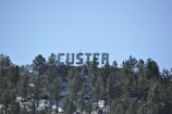 Large letters spelling 'CUSTER' are mounted on a hillside covered with pine trees. The letters are white and are positioned against a clear blue sky, providing a stark contrast to the dense green foliage.
