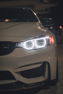 A close-up of a white car's front end, featuring sleek headlights that are illuminated with LED lights, creating a modern and stylish appearance. The car's polished surface reflects light subtly in the dark atmosphere.