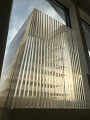 A structure made of clear, parallel, vertical plastic or glass panels, positioned against a background of a softly lit window. The light creates a play of shadows and reflections across the surface.