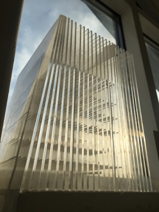 A structure made of clear, parallel, vertical plastic or glass panels, positioned against a background of a softly lit window. The light creates a play of shadows and reflections across the surface.