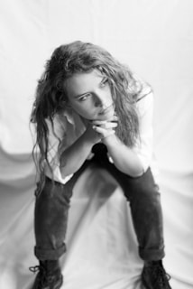 A person with long hair is seated, looking contemplatively to the side. The setting is minimalist, featuring a light-colored backdrop. The individual is wearing a light shirt and dark pants, with their hands clasped under their chin.