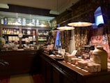 A vintage-style shop interior with dark wood furnishings and ornate floral curtains. It features shelves filled with jars, bottles, and boxes, showcasing a variety of products. A mannequin dressed in historical attire is behind the counter. Warm lighting from hanging lamps creates a cozy atmosphere.