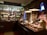 A vintage-style shop interior with dark wood furnishings and ornate floral curtains. It features shelves filled with jars, bottles, and boxes, showcasing a variety of products. A mannequin dressed in historical attire is behind the counter. Warm lighting from hanging lamps creates a cozy atmosphere.