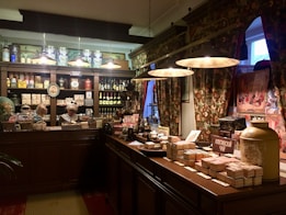 A vintage-style shop interior with dark wood furnishings and ornate floral curtains. It features shelves filled with jars, bottles, and boxes, showcasing a variety of products. A mannequin dressed in historical attire is behind the counter. Warm lighting from hanging lamps creates a cozy atmosphere.