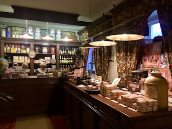 A vintage-style shop interior with dark wood furnishings and ornate floral curtains. It features shelves filled with jars, bottles, and boxes, showcasing a variety of products. A mannequin dressed in historical attire is behind the counter. Warm lighting from hanging lamps creates a cozy atmosphere.