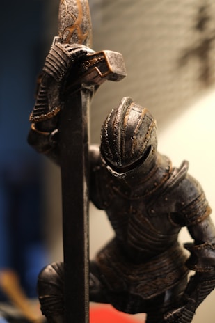 A medieval knight wearing ornate, dark metal armor kneels with one hand resting on a large, elaborate sword. The background is blurred, creating a contrast that draws attention to the detailed textures and design of the knight's armor and weapon. The lighting highlights the intricate patterns and aged appearance of the metal.