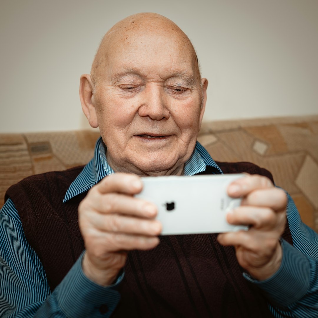 Portrait of nice cheerful positive cheery stylish old man sitting on divan holding in hands a smartphone 