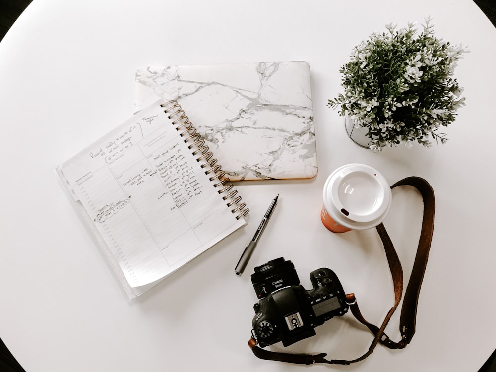 black nikon dslr camera on white notebook beside white ceramic mug