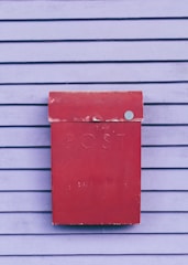 A weathered red postbox is mounted on a wall with horizontal wooden slats painted in a soft purple color. The word 'POST' is embossed on the front of the postbox.