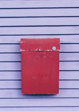 A weathered red postbox is mounted on a wall with horizontal wooden slats painted in a soft purple color. The word 'POST' is embossed on the front of the postbox.
