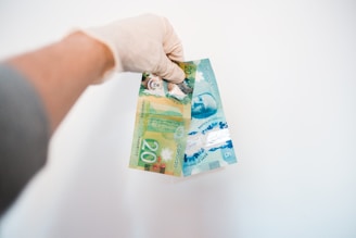 A hand wearing a glove is holding Canadian banknotes against a plain white background. The banknotes are vibrant and colorful, primarily featuring a green and blue design, with the denominations clearly visible as 20 and 5.