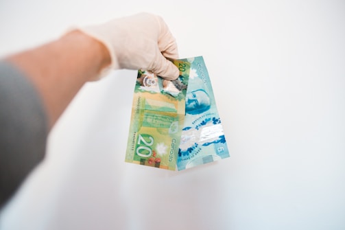 A hand wearing a glove is holding Canadian banknotes against a plain white background. The banknotes are vibrant and colorful, primarily featuring a green and blue design, with the denominations clearly visible as 20 and 5.
