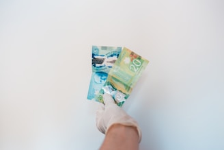 A gloved hand is holding Canadian banknotes against a plain white background. The banknotes include a five-dollar bill and a twenty-dollar bill, both with colorful designs.