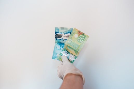 A gloved hand is holding Canadian banknotes against a plain white background. The banknotes include a five-dollar bill and a twenty-dollar bill, both with colorful designs.