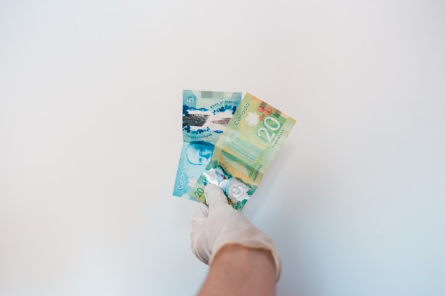 A gloved hand is holding Canadian banknotes against a plain white background. The banknotes include a five-dollar bill and a twenty-dollar bill, both with colorful designs.
