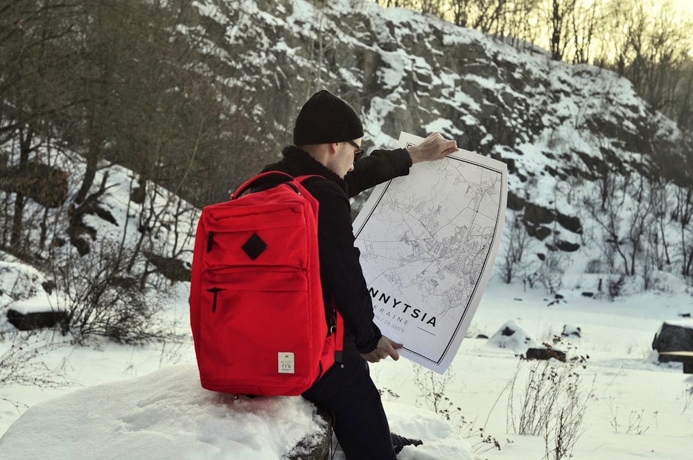 uomo in giacca nera che trasporta zaino rosso e nero in piedi su terreno coperto di neve durante il giorno