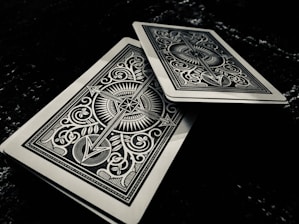 Two intricately designed playing cards feature symmetrical patterns with sharp lines and ornate symbols. One card is slightly offset above the other on a dark surface, adding depth to the composition.
