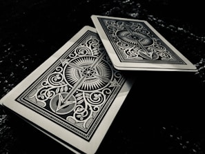 Two intricately designed playing cards feature symmetrical patterns with sharp lines and ornate symbols. One card is slightly offset above the other on a dark surface, adding depth to the composition.
