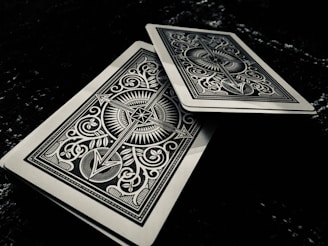 Two intricately designed playing cards feature symmetrical patterns with sharp lines and ornate symbols. One card is slightly offset above the other on a dark surface, adding depth to the composition.