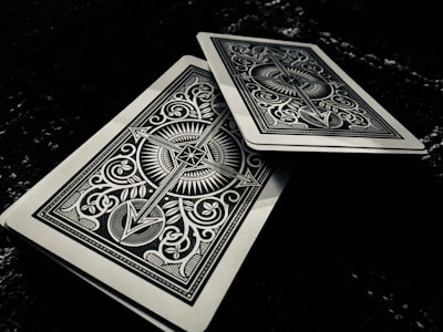 Two intricately designed playing cards feature symmetrical patterns with sharp lines and ornate symbols. One card is slightly offset above the other on a dark surface, adding depth to the composition.