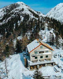 A rustic house with a snow-covered roof is nestled among snow-laden trees and mountainous terrain. The structure is built with stone and wood, featuring solar panels on the roof. The surrounding landscape consists of densely packed pine trees and towering snowy peaks under a clear blue sky.
