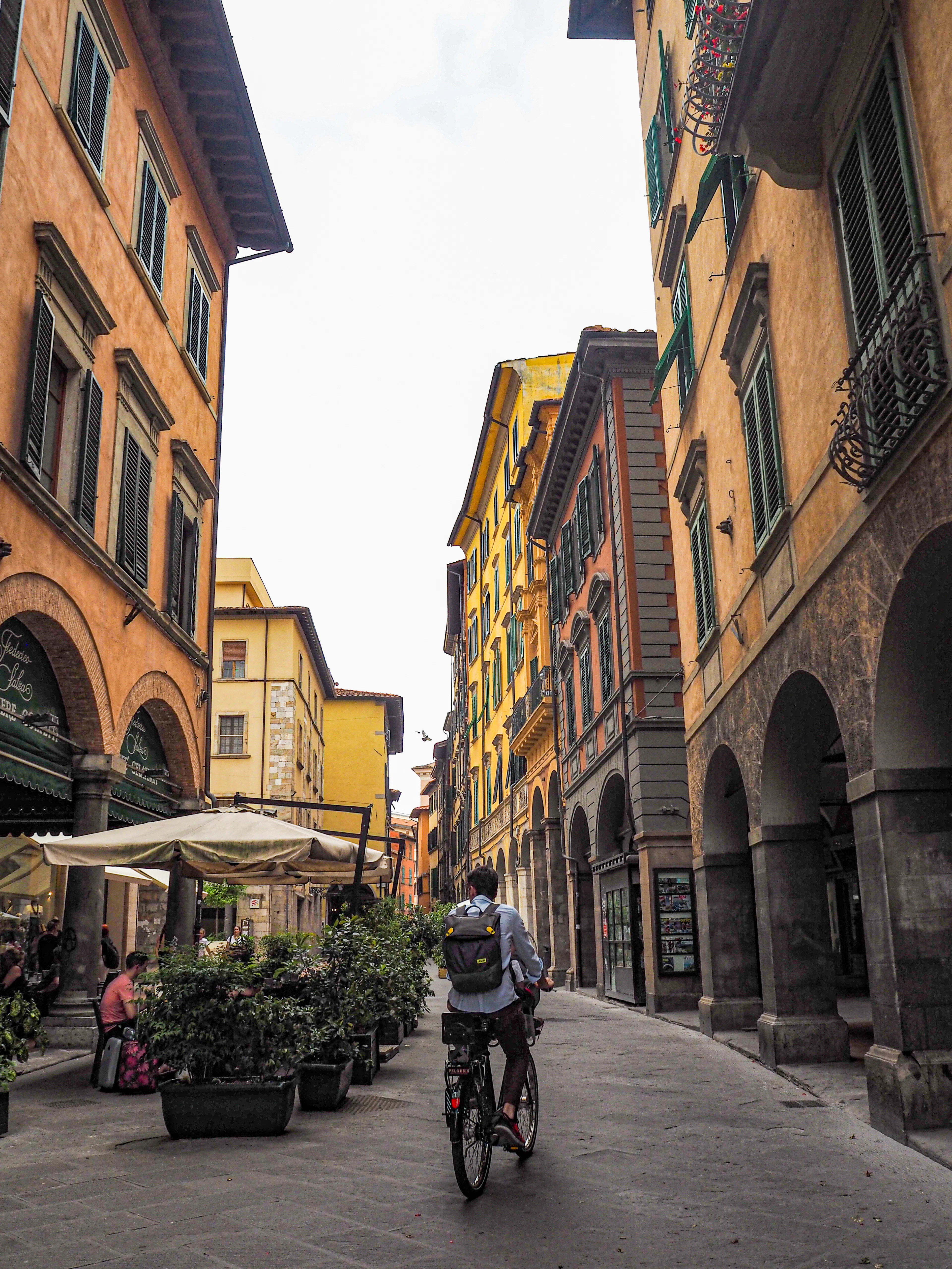 A cyclist riding down Pisa's arcaded old-town shopping street, between ochre palazzi and cafe awnings