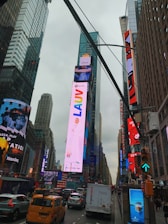 A bustling urban scene featuring a busy street with yellow cabs, various vehicles, and towering skyscrapers. Bright digital billboards display colorful advertisements, making the area vibrant and lively.
