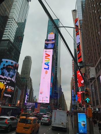 A bustling urban scene featuring a busy street with yellow cabs, various vehicles, and towering skyscrapers. Bright digital billboards display colorful advertisements, making the area vibrant and lively.