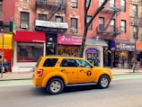 A yellow taxi is parked on a city street, in front of a row of brick buildings with various storefronts including a clothing shop and a cleaners. The street has a marked bike lane. There are signs for rentals in the windows above the shops, and a few pedestrians are visible.