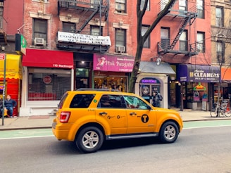 A yellow taxi is parked on a city street, in front of a row of brick buildings with various storefronts including a clothing shop and a cleaners. The street has a marked bike lane. There are signs for rentals in the windows above the shops, and a few pedestrians are visible.
