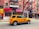 A yellow taxi is parked on a city street, in front of a row of brick buildings with various storefronts including a clothing shop and a cleaners. The street has a marked bike lane. There are signs for rentals in the windows above the shops, and a few pedestrians are visible.
