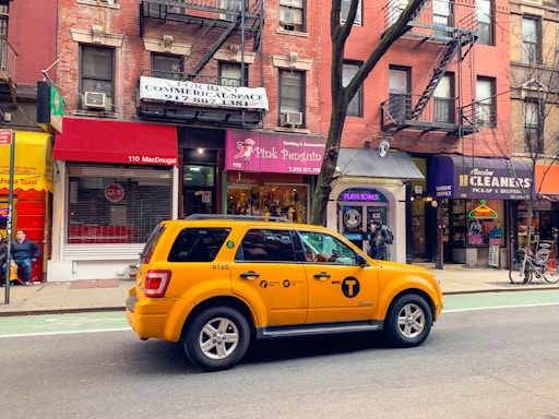 A yellow taxi is parked on a city street, in front of a row of brick buildings with various storefronts including a clothing shop and a cleaners. The street has a marked bike lane. There are signs for rentals in the windows above the shops, and a few pedestrians are visible.