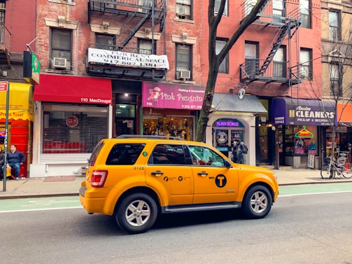 A yellow taxi is parked on a city street, in front of a row of brick buildings with various storefronts including a clothing shop and a cleaners. The street has a marked bike lane. There are signs for rentals in the windows above the shops, and a few pedestrians are visible.
