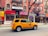 A yellow taxi is parked on a city street, in front of a row of brick buildings with various storefronts including a clothing shop and a cleaners. The street has a marked bike lane. There are signs for rentals in the windows above the shops, and a few pedestrians are visible.