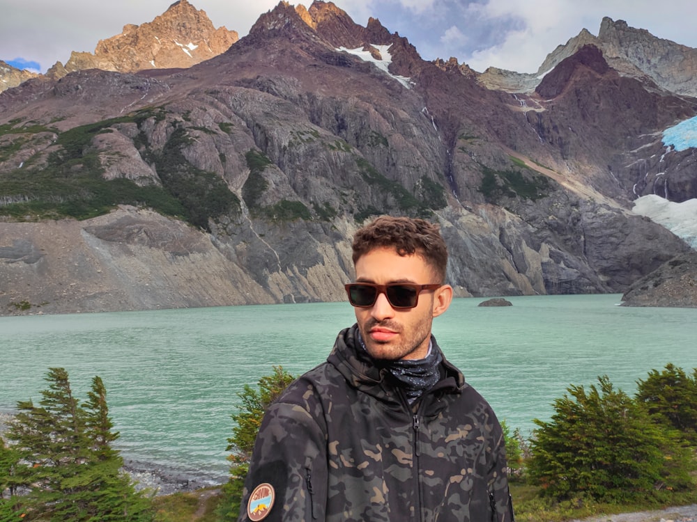 man in black and gray camouflage jacket wearing black sunglasses standing near body of water during