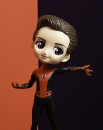 A figurine of a character with large eyes, wearing a red and black costume reminiscent of a superhero outfit. The background is split into two contrasting colors: orange and dark purple.