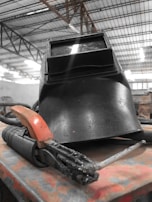 A black welding helmet and a welding torch rest on a flat surface in an industrial setting. The background features metal beams and a high ceiling, indicating a workshop or factory environment.