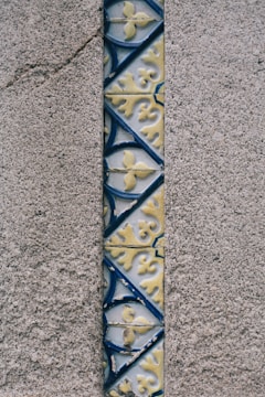 A narrow vertical strip of ornate ceramic tiles in shades of blue and yellow is embedded in a rough, textured gray concrete wall. The tiles feature intricate floral patterns and geometric shapes.